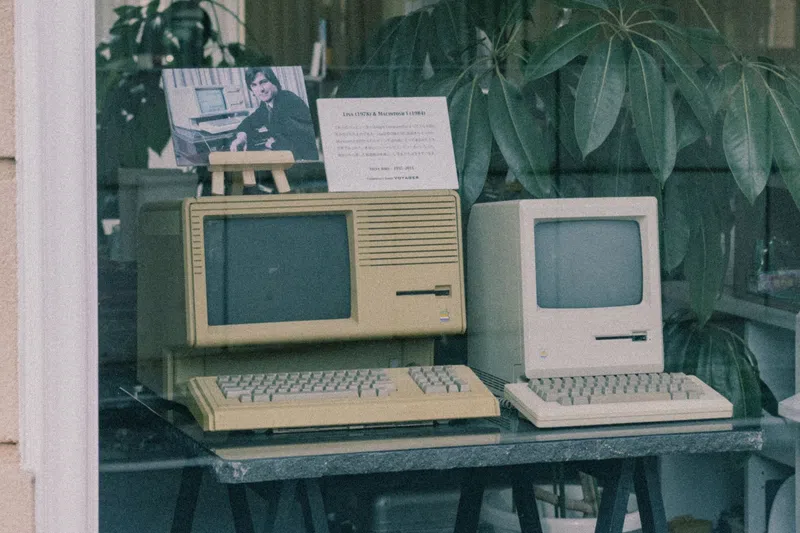 Two vintage personal computers displayed on a table, one beige and one cream, with a photo and plant background.