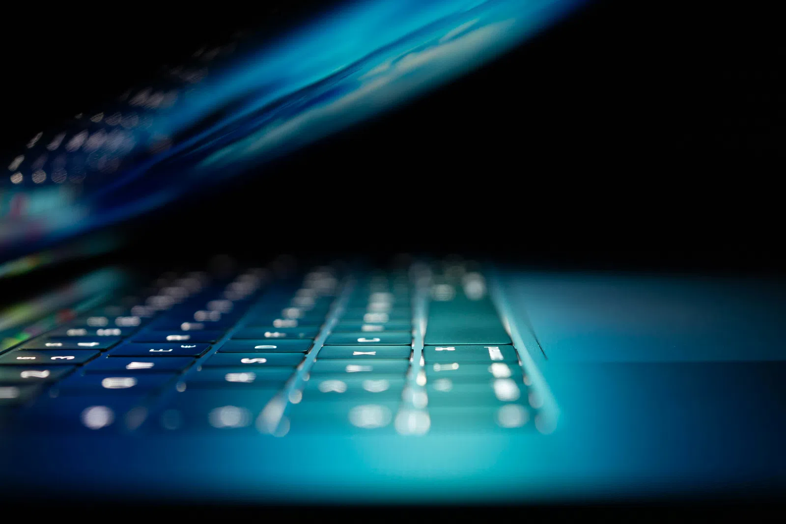 Close-up of a laptop keyboard with blue backlighting in a dark environment highlighting the keys and screen edge.